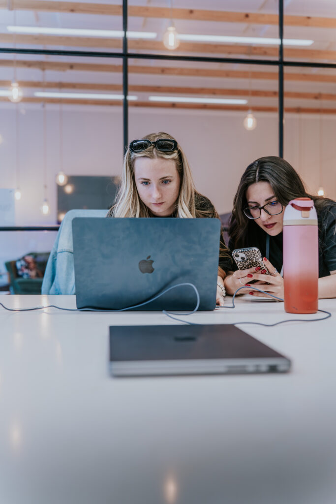 Two women sit at a table, one using a laptop and the other looking at a phone.