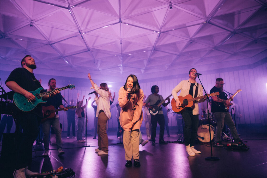A band of eight people playing guitars, singing, and dancing during a live performance.