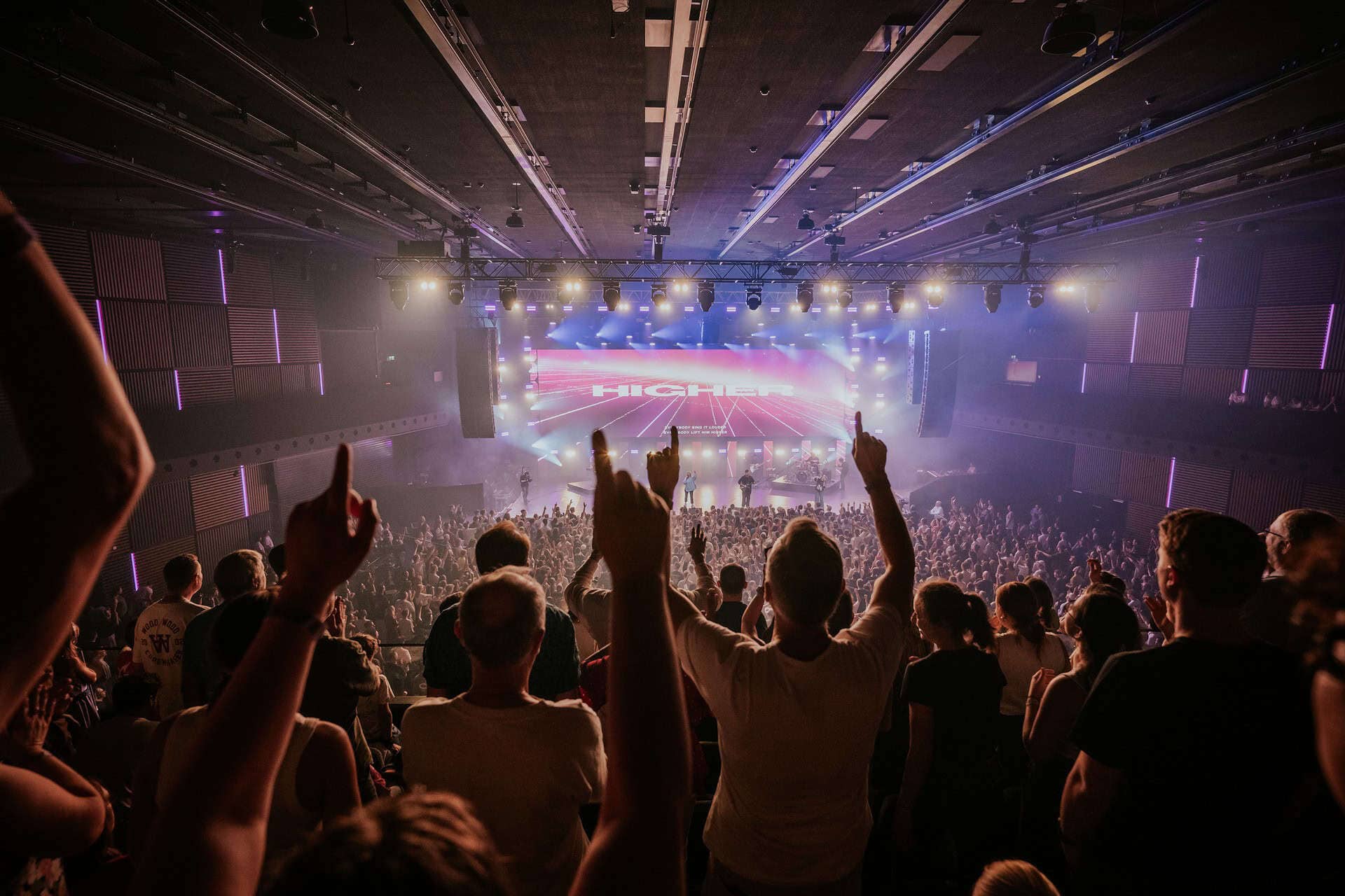 Audience in a concert hall facing a brightly lit stage with colorful lights.