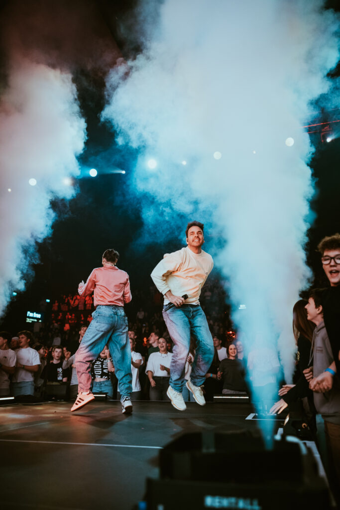 An image of two men jumping on stage with smoke effects in the background.