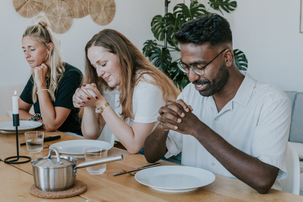 Three people praying at a dining table with plates, glasses, and a pot.