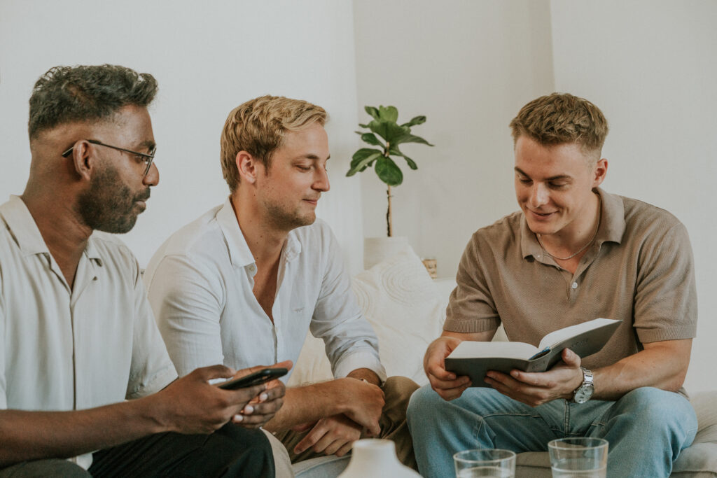 Three men sitting on a sofa, engaged in a discussion, with one reading a book.