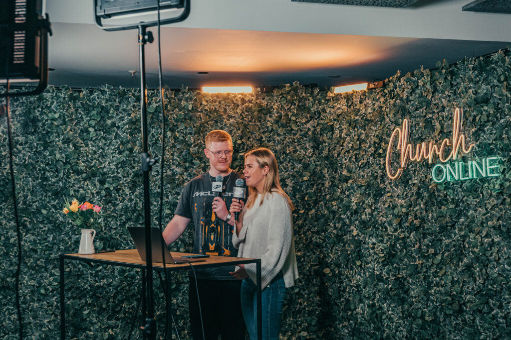Two people speaking into microphones in front of a green leafy wall with a neon sign that reads "Chu.