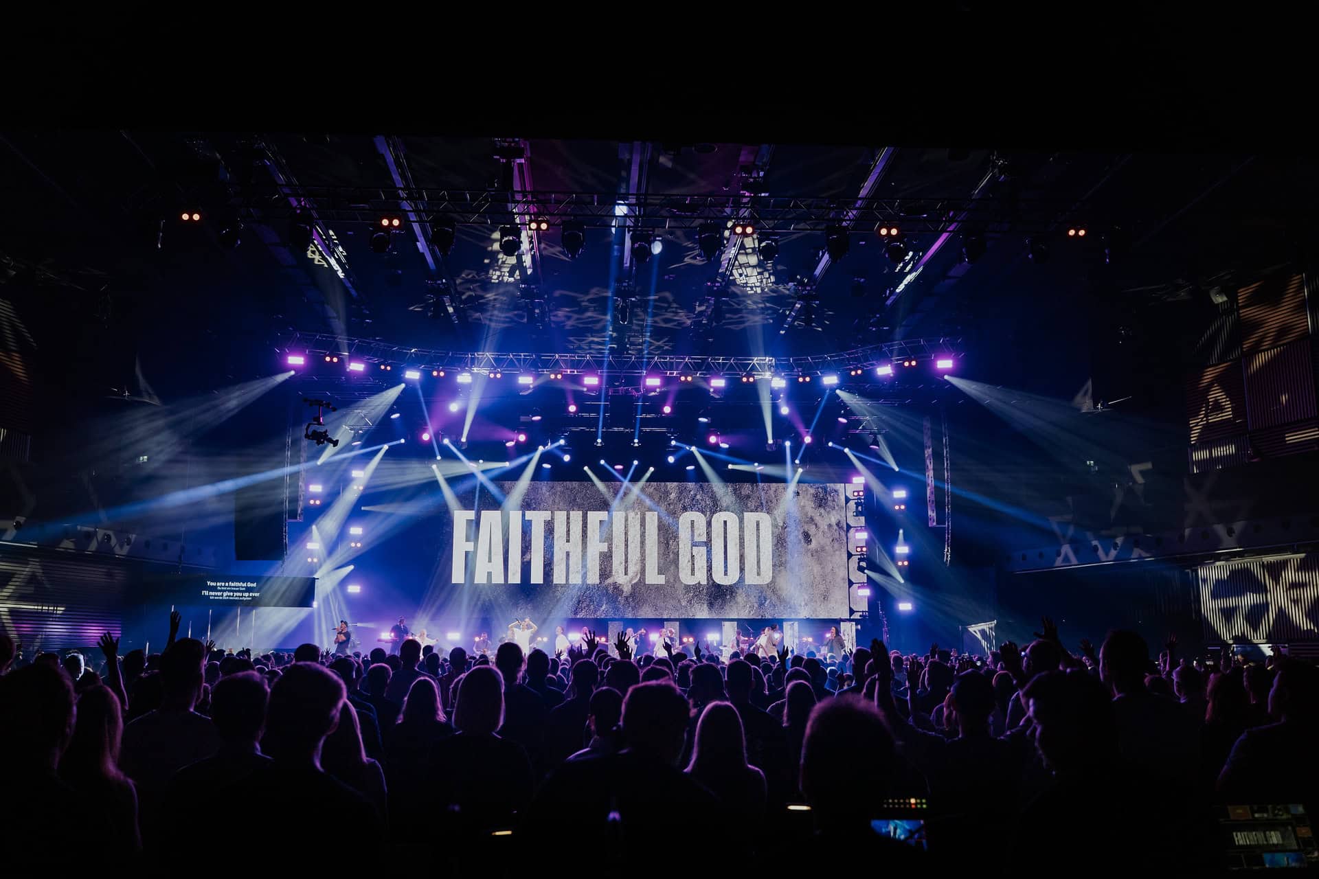 Hochglanzlichtaufnahme eines modernen Konzertraumes mit buntem Licht- und Laser-Show, Bühne mit der Aufschrift „FAITHFUL GOD“, voll besetztes Publikum, geeignet für christliche Veranstaltungen und spirituelle Konzerte.