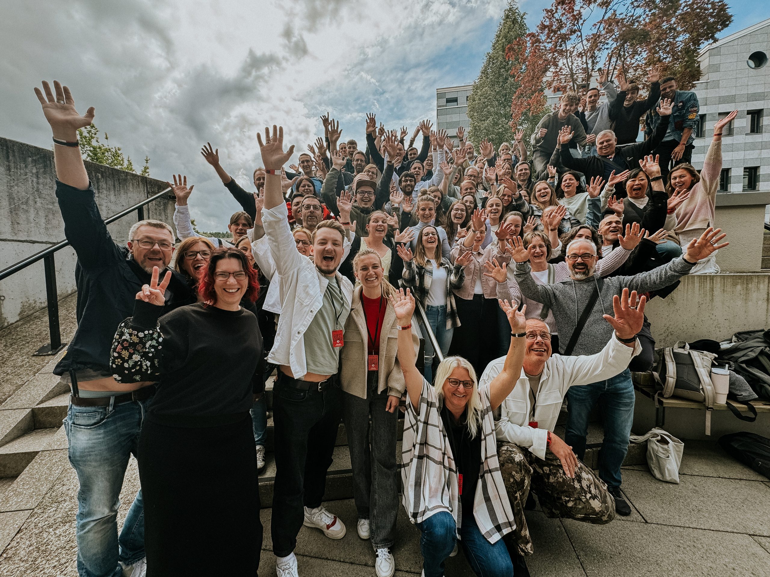 Viele Menschen stehen auf einer Treppe im Freien, heben die Hände und lächeln.