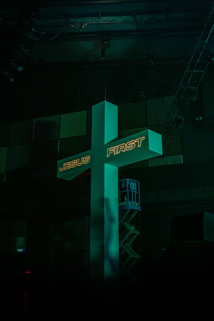 Gekreuztes, leuchtendes Kreuz mit der Aufschrift "Jesus First" in einer modernen Kirche oder Veranstaltungsraum, symbolisiert christlichen Glauben und Priorität Jesu in einer inspirierenden Atmosphäre.