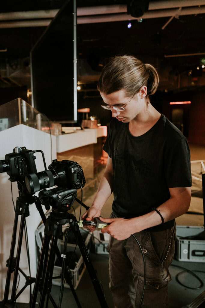 Moderner Kameramann stellt Kameraeinstellungen ein bei Filmdreh in einem dunklen Studio, Fokus auf Filmproduktion, Videokunst und kreative Medienarbeit, professionelles Filmemachen.