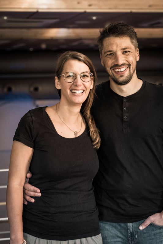 Zwei Menschen, eine Frau und ein Mann, lächeln in einem Raum.