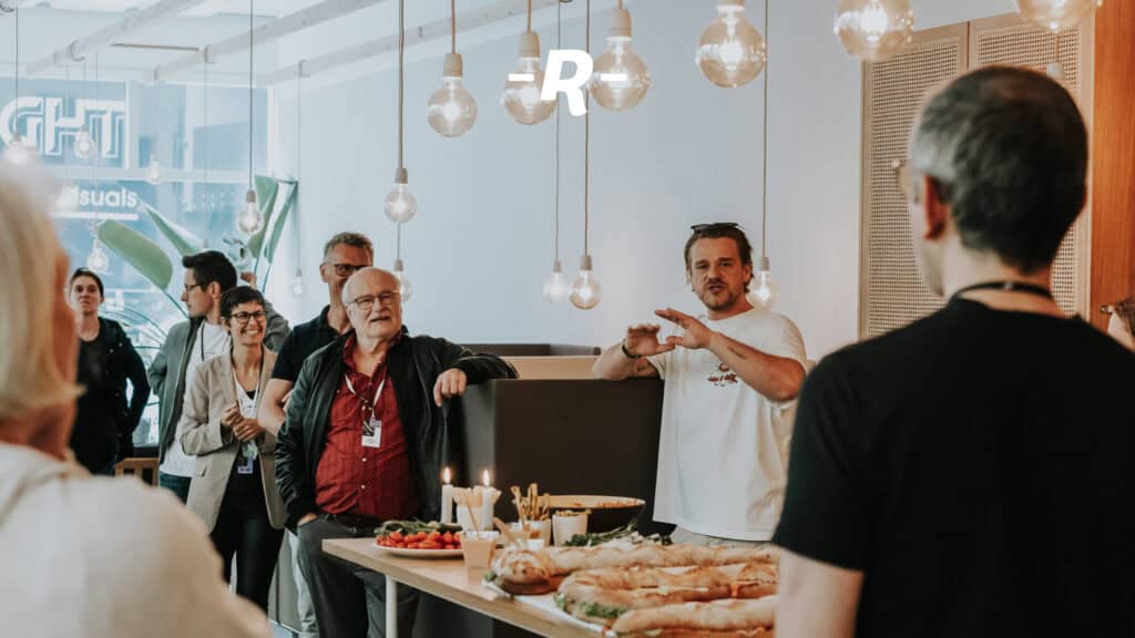 Diversity of people enjoying a communal meal in a modern, bright room with decorative light bulbs and wood paneling; expression of cohesion and social interaction in an informal setting.