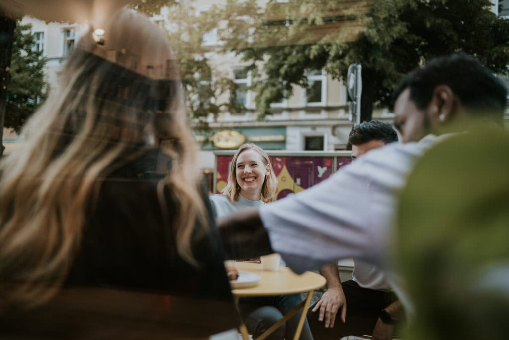 Freundliche junge Menschen beim Treffen im öffentlichen Raum, lächelnde Frau im Mittelpunkt, lockere Atmosphäre, urbanes Umfeld, Begegnung der Jugendlichen, soziale Interaktion, fröhliches Beisammensein, Jugendarbeit, Gemeinschaftsgefühl, Stadtleben, junge Erwachsenen, Teens, Outdoor-Event, Freundschaft, positive Stimmung, Gemeinschaft.