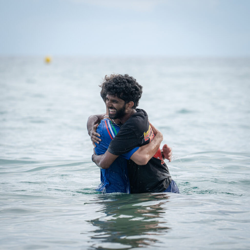 Zwei Männer umarmen sich im Wasser, lächelnd.