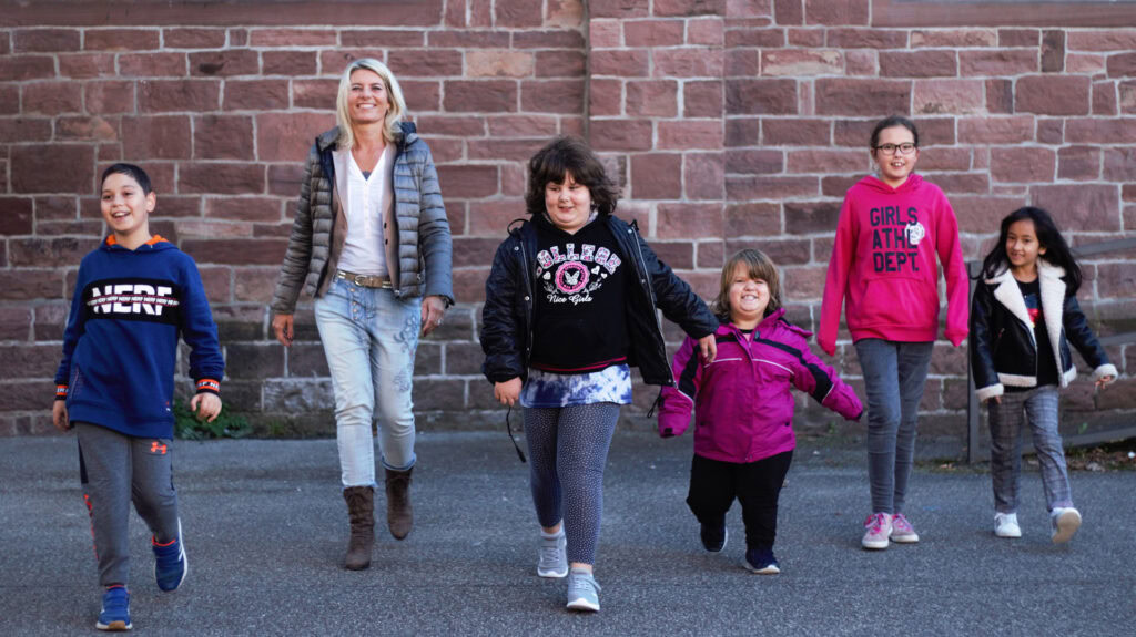 Fünf Kinder und eine erwachsene Frau gehen an einer Ziegelmauer entlang.
