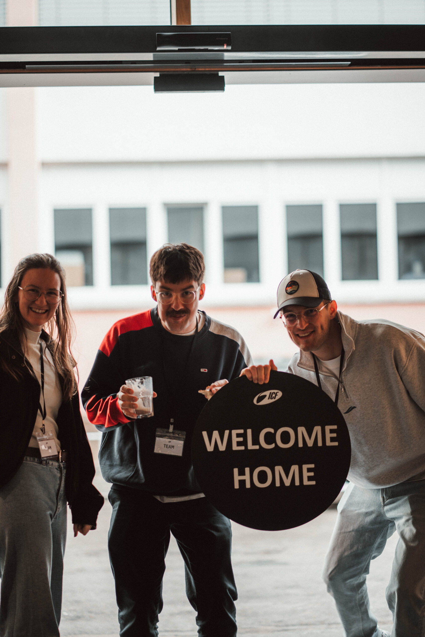 Drei Menschen begrüßen sich herzlich bei einer Veranstaltung im ICF Schwarzwald Bodensee, halten ein "Welcome Home" Schild, symbolisieren Gemeinschaft und Gastfreundschaft.