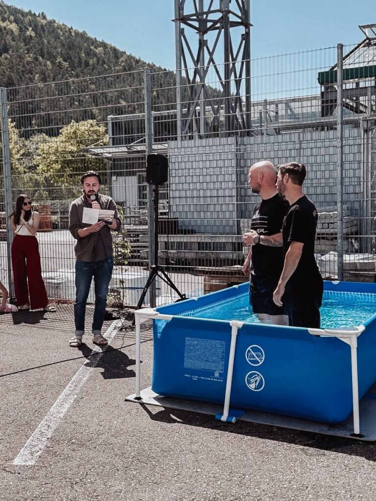 Zwei Männer werden bei einer Taufe im Außenbecken des ICF Schwarzwald Bodensee getauft, während eine Frau die Zeremonie filmt. Das Event findet auf einem Parkplatz mit Industrieanlagen im Hintergrund.