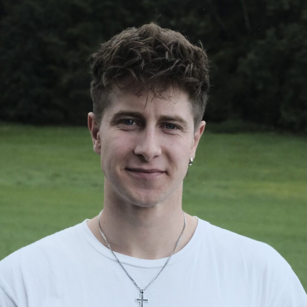 Junger Mann mit Kurzhaarfrisur, weißem T-Shirt, silbernem Kreuz-Halskette und Ohrring, steht im Freien auf grüner Wiese, freundliches Lächeln, natürliche Umgebung, moderne christliche Kleidung, ernsthafter Ausdruck.