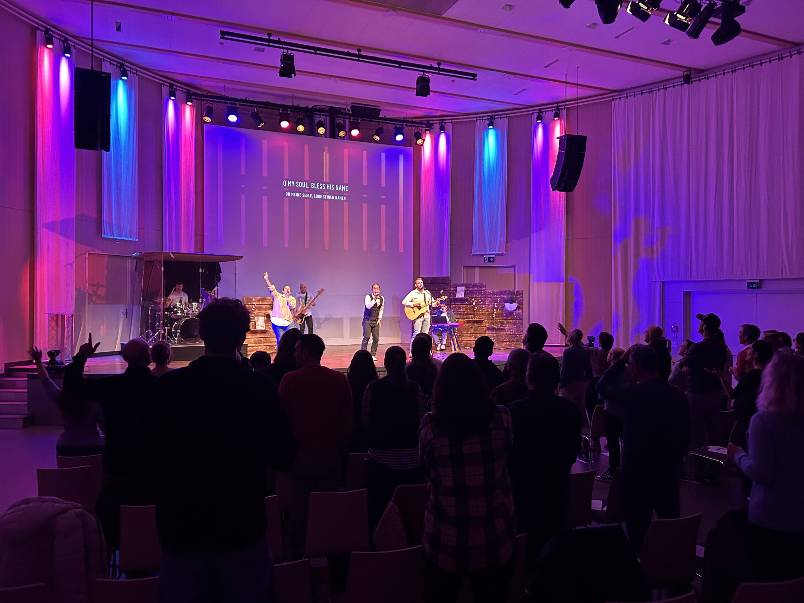 Bühne mit Musikern, Publikum im Saal, bunte Lichtbeleuchtung, Projektor an der Wand.