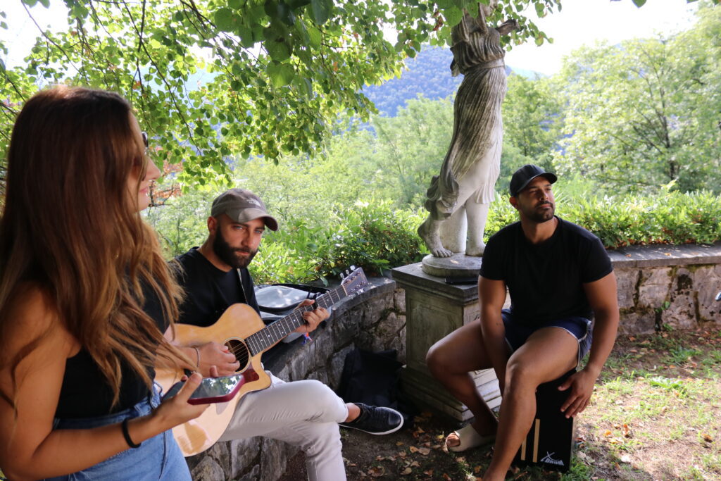 Tre persone suonano e ascoltano musica in un parco con alberi e statue.