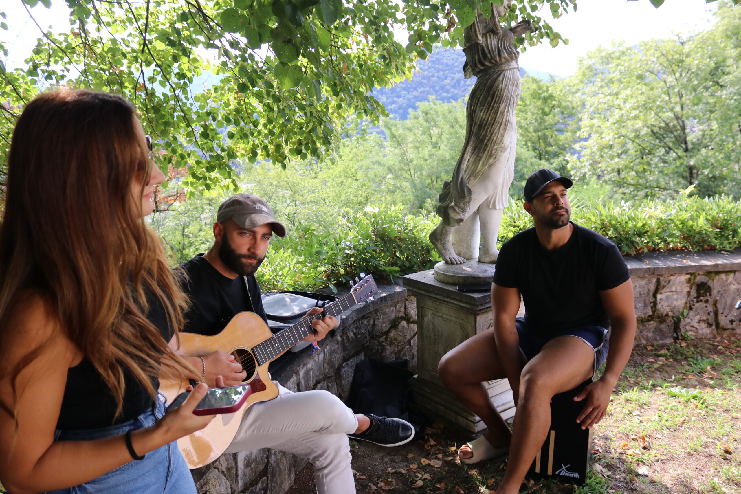 Tre persone suonano e ascoltano musica in un parco con alberi e statue.