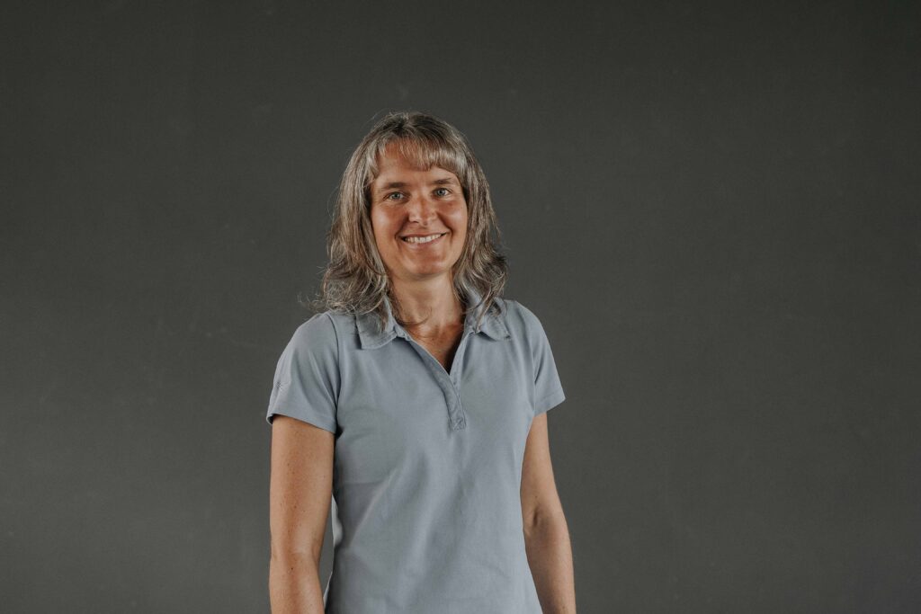 Frau mit Schulterlänge, grauem Haar, lächelnd, in hellblauem Polo-Shirt vor grauem Hintergrund.