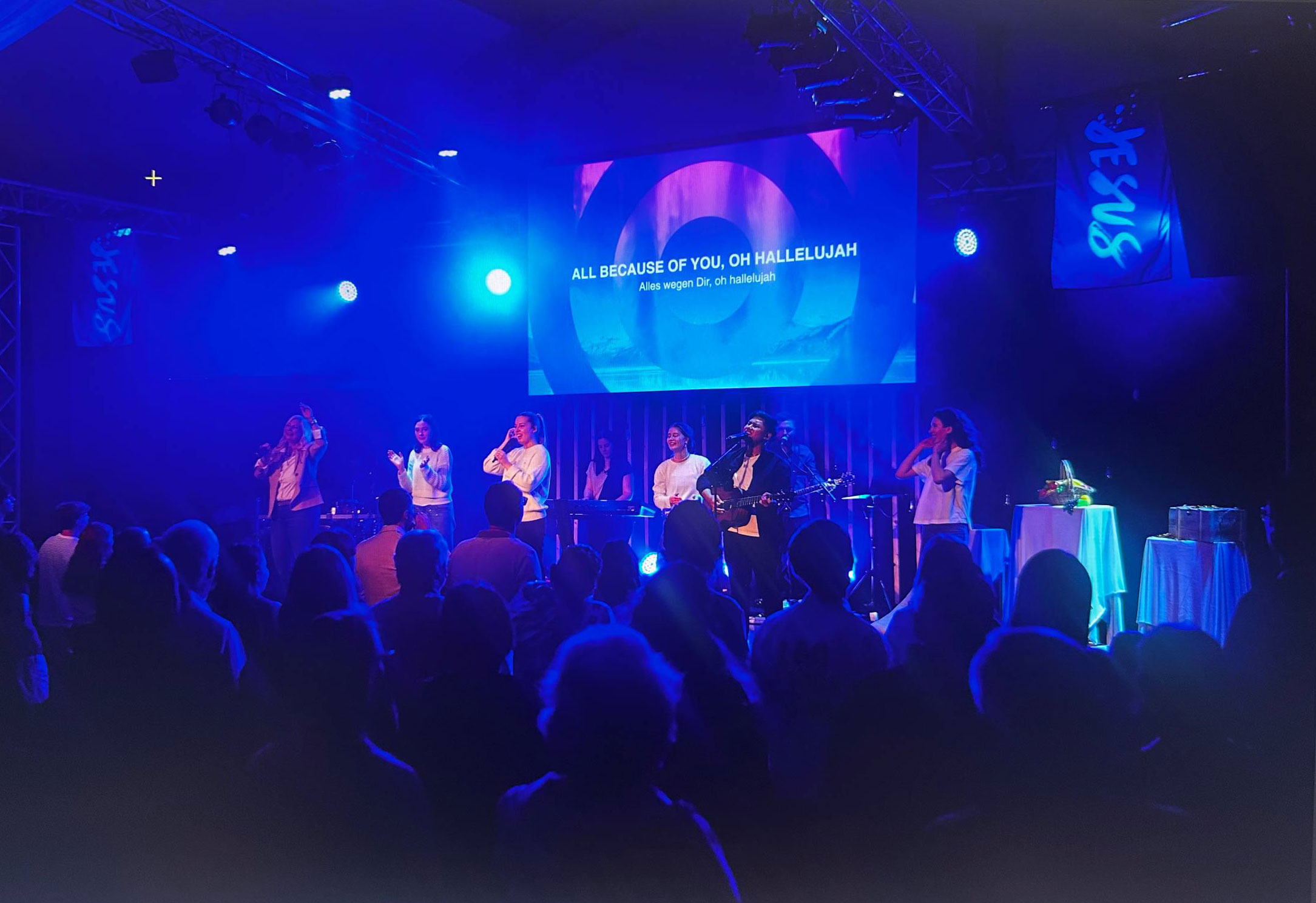 Menschen auf einer Bühne beim Gottesdienst mit Musik, im dunklen Raum mit blauen Lichtern.