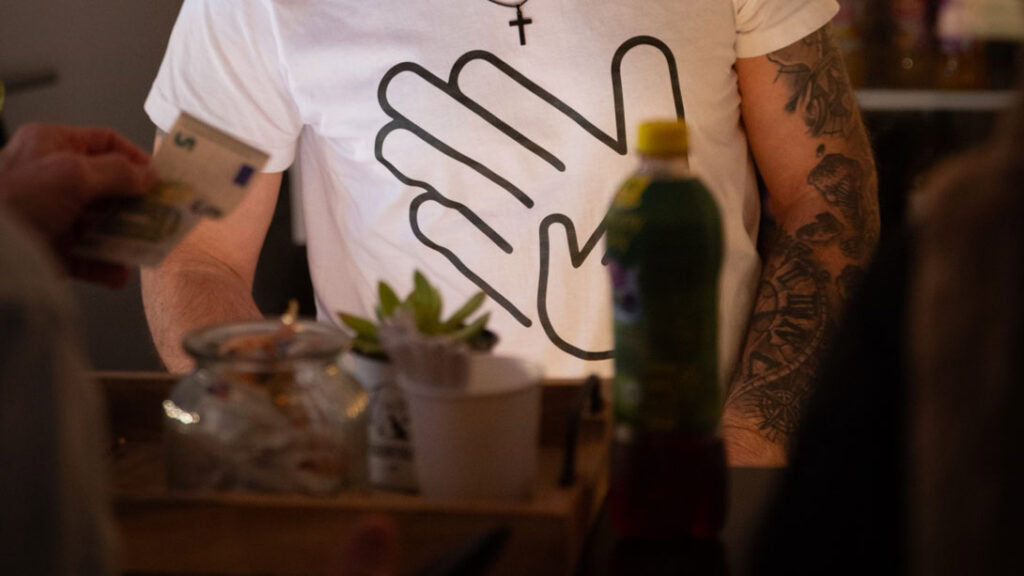 Mensch mit Tattoo auf dem linken Arm, trägt weißes T-Shirt mit Hand-Logo.