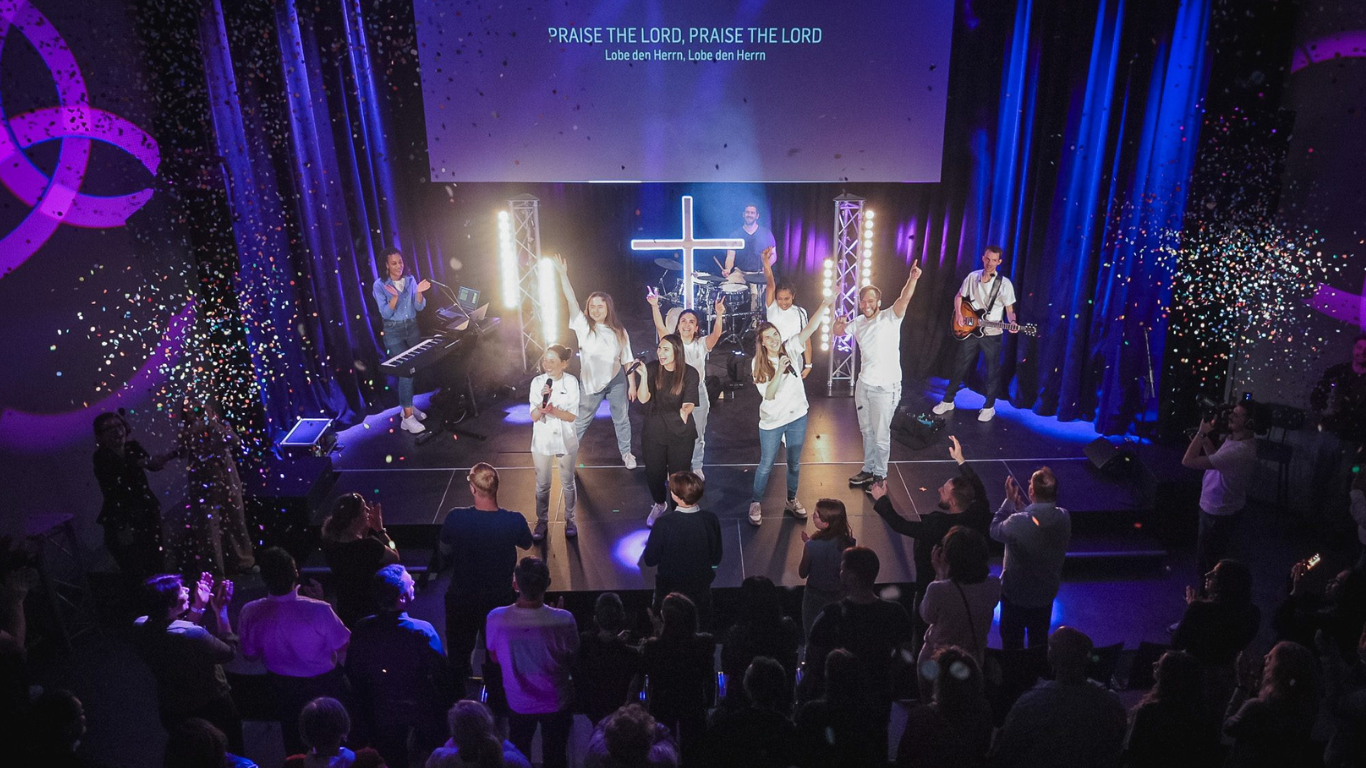 Menschen auf einer Bühne während eines Gottesdienstes, mit Musikern und Sängern, im Hintergrund ein.