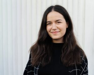 Junge Frau mit langen braunen Haaren, die vor einer weißen Wand steht und lächelt.