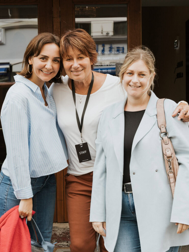 Drei Frauen stehen draußen vor einem Gebäude, lächeln und umarmen sich.