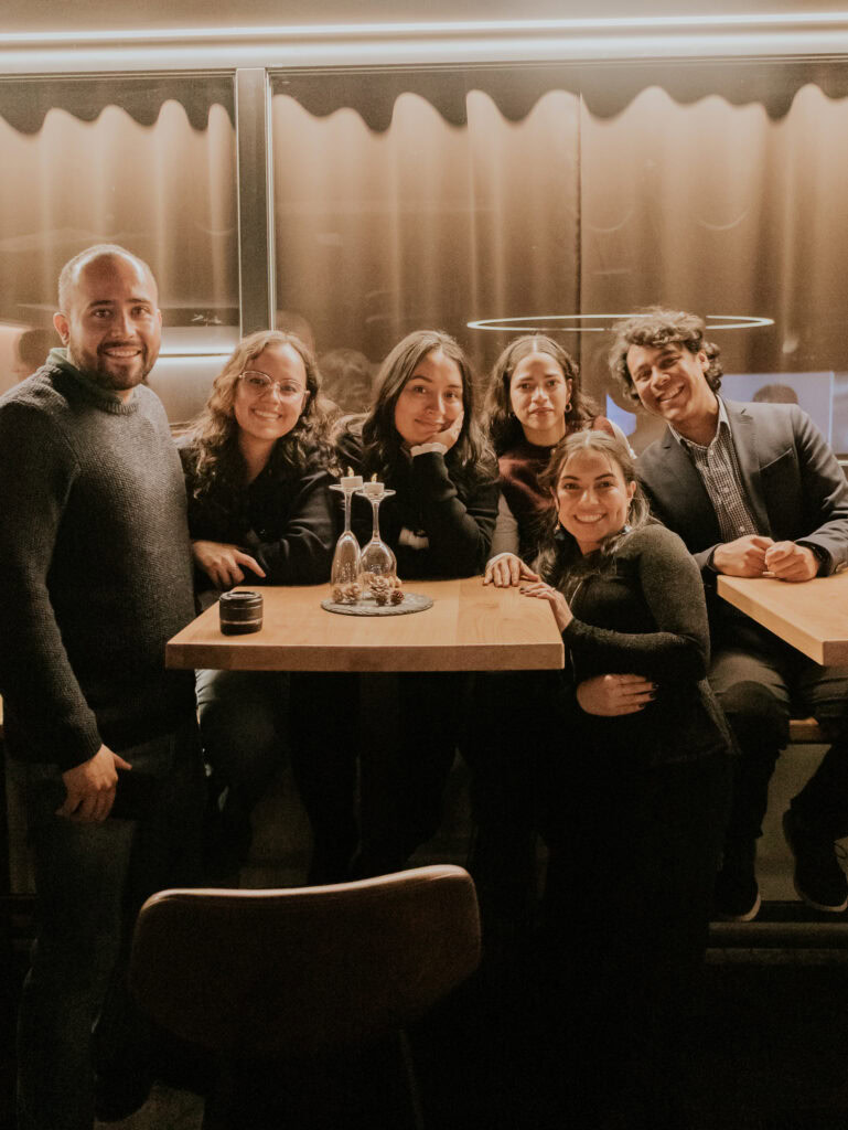 Sechs Menschen stehen um einen Tisch, lachen und posieren für die Kamera.