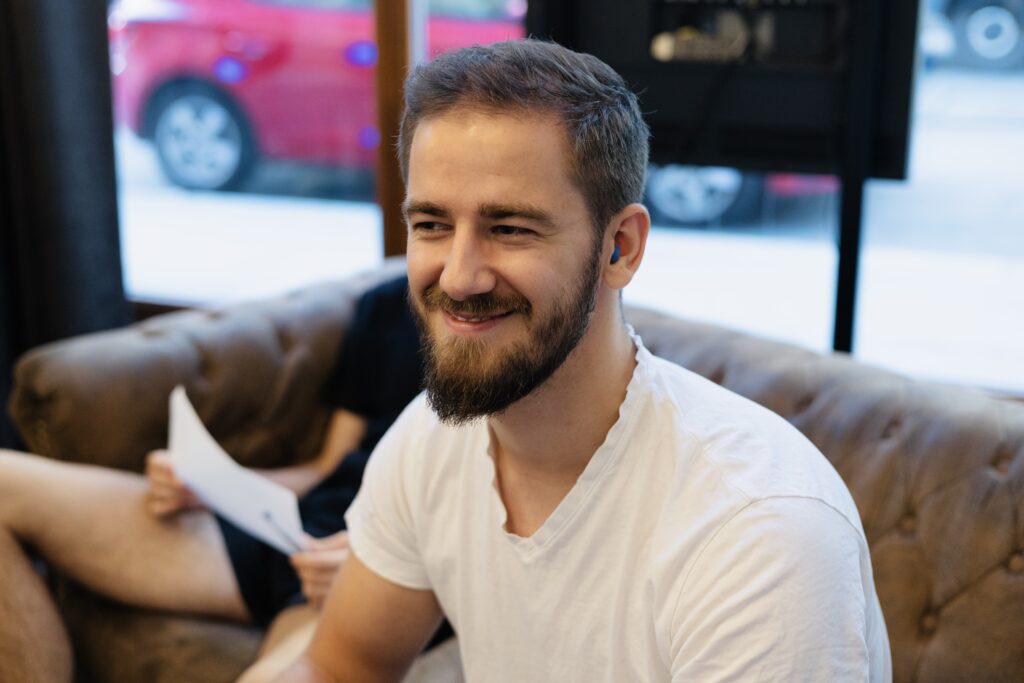 Mann mit Bart und weißen T-Shirt sitzt auf einer Couch, im Hintergrund eine Person mit Papier.