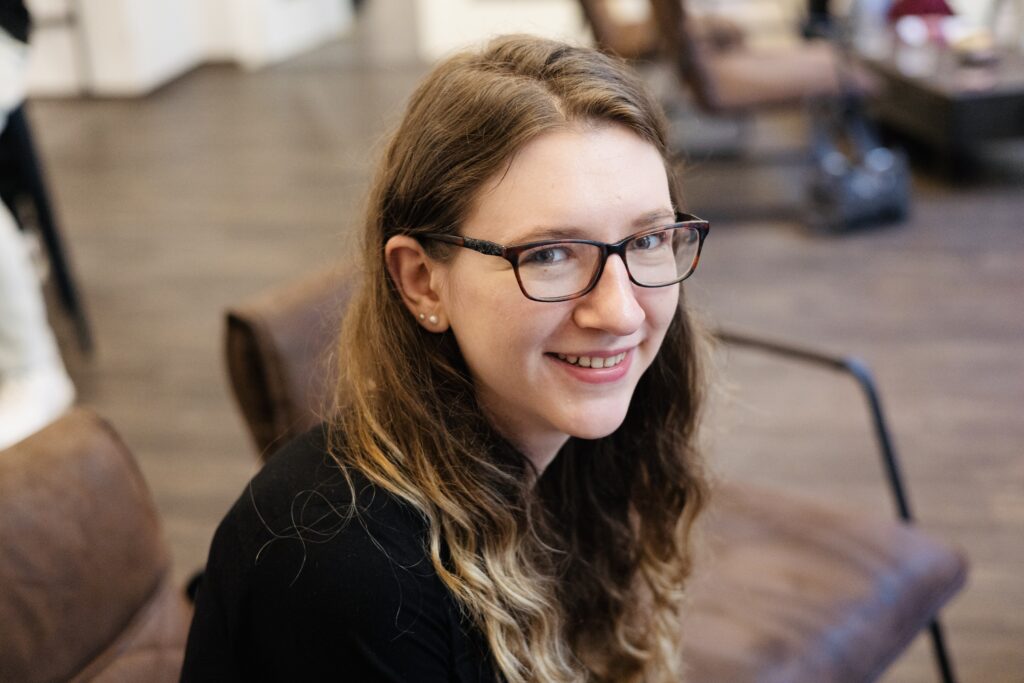 Junge frau mit brille, lächelnd, sitzt in einem Raum mit Holzboden und Möbeln.