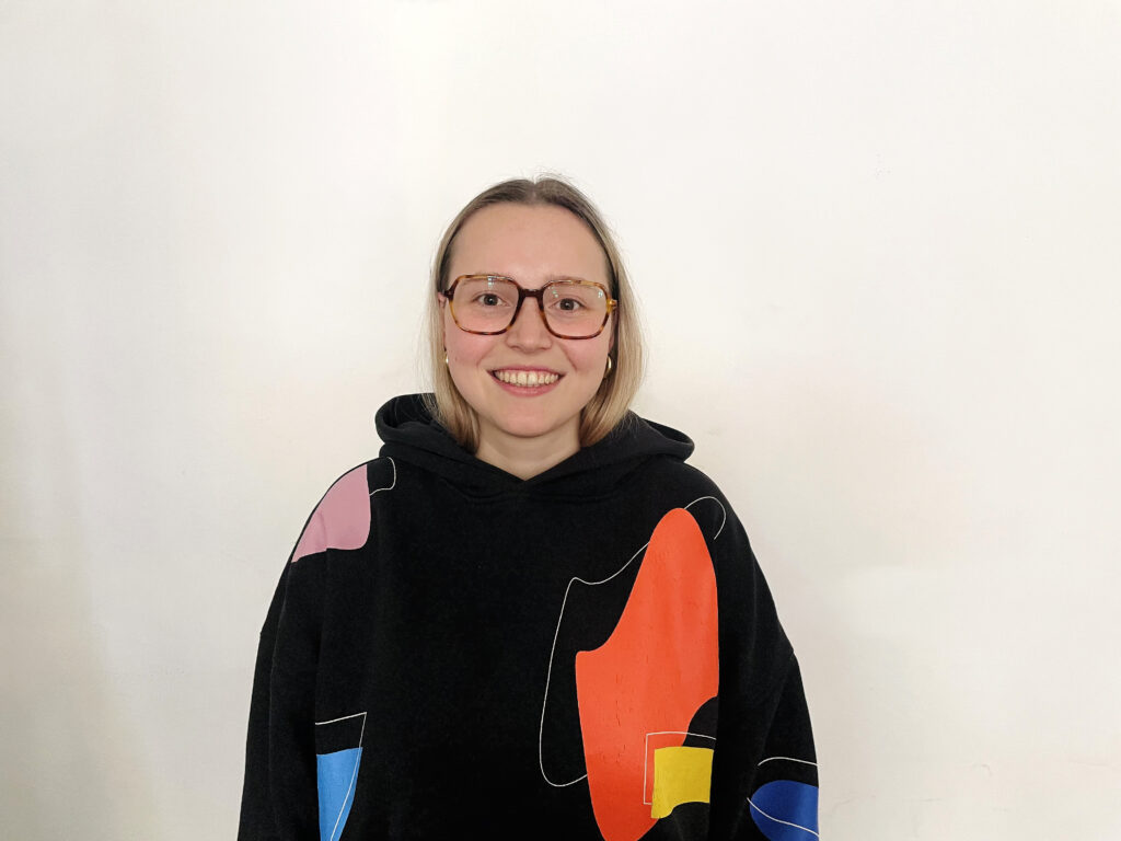 Junge Frau mit Brille trägt schwarzen Hoodie mit buntem Logo, lächelt.