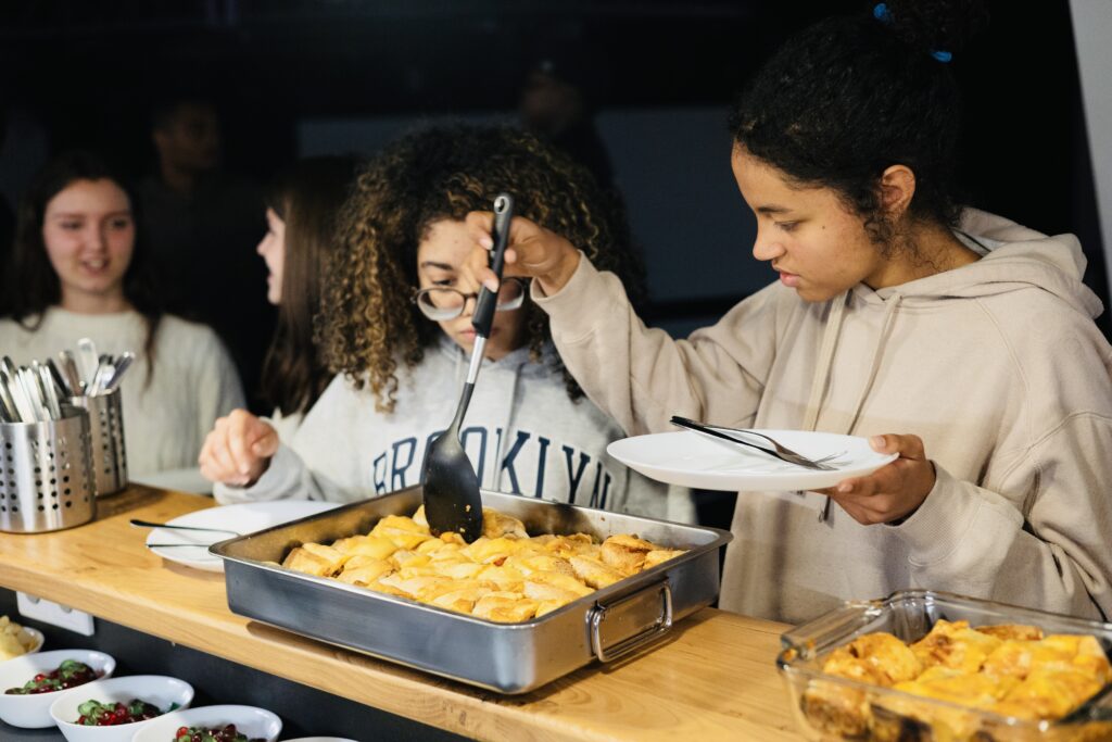 Zwei junge Frauen bedienen sich an einem Buffet mit Kartoffeln, eine schaufelt Kartoffeln auf ihren.