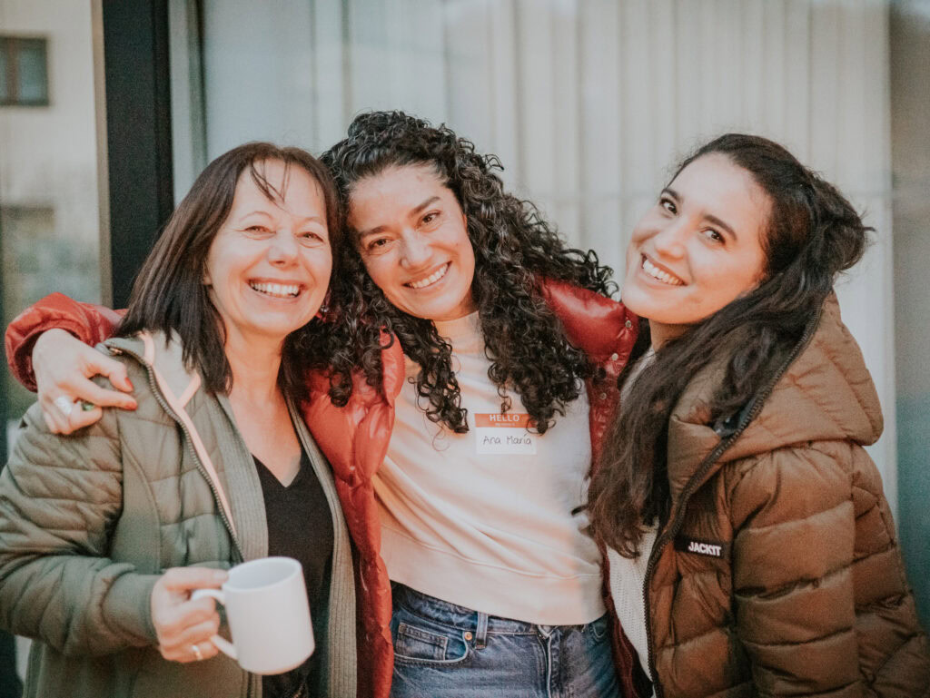 Drei Frauen stehen zusammen, lächeln und umarmen sich, eine hält eine Tasse.