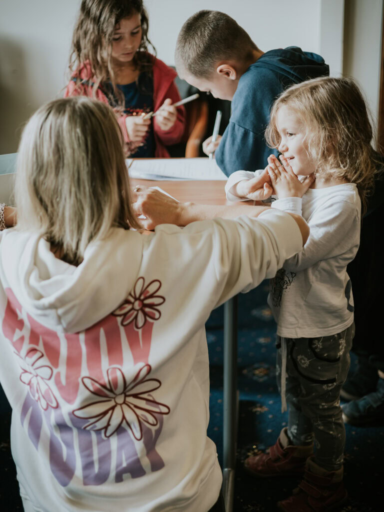 Zwei Kinder werden von einer Erwachsenen bei den Hausaufgaben unterstützt, während andere Kinder im.