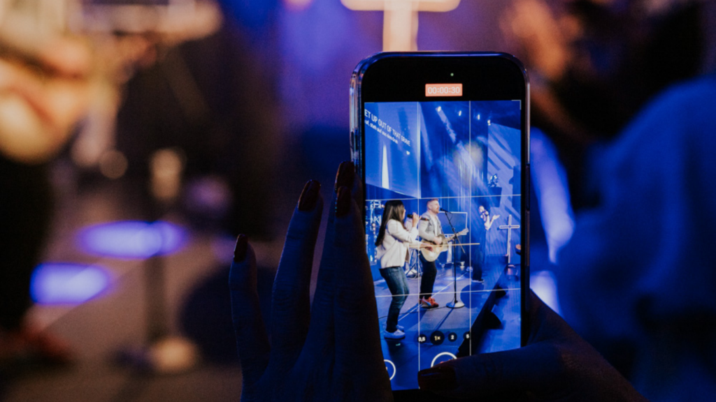 Person hält Smartphone hoch, zeigt einen Live-Musikauftritt mit mehreren Musikern auf der Bühne.