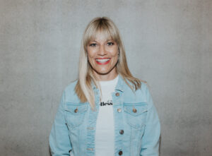 Lächelnde Frau mit blonden Haaren und Jeansjacke vor grauem Hintergrund.