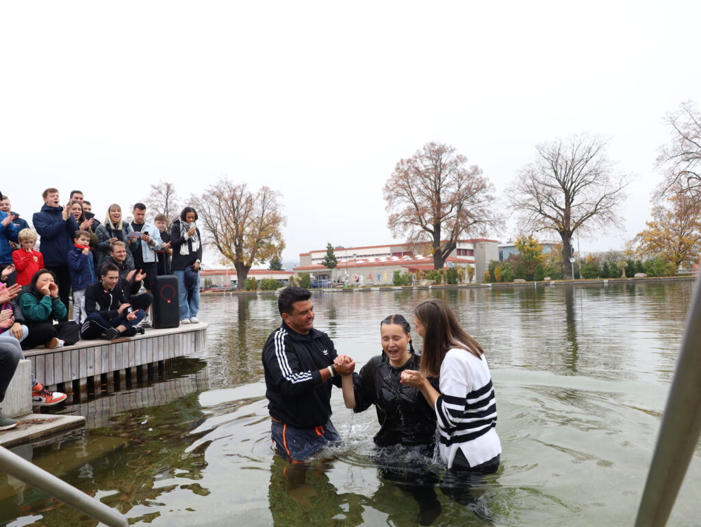 Menschen beim Taufritual im Wasser, umgeben von Zuschauern am Ufer.