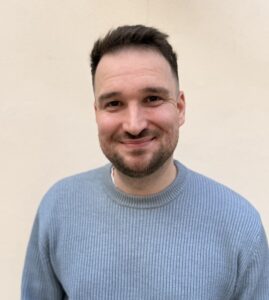 Mann mit dunklem Haar und Bart, trägt blauen Pullover, lächelt.