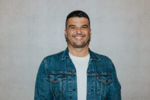 Mann mit dunklem Haar und Bart trägt Denimjacke und weißes T-Shirt.