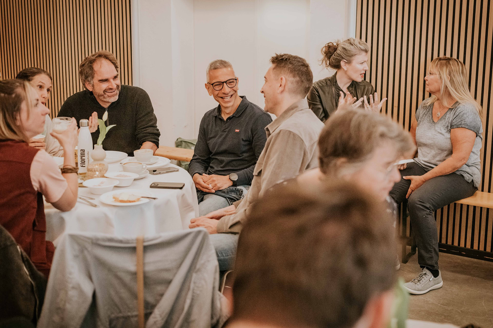 Menschen sitzen um einen runden Tisch, trinken Kaffee und unterhalten sich.