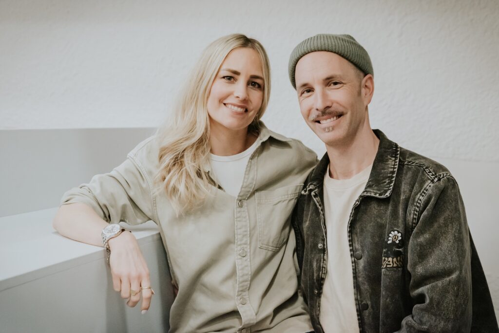 Ein lachendes Paar steht vor einer weißen Wand, die Frau hat blonde Haare, der Mann trägt einen grau.