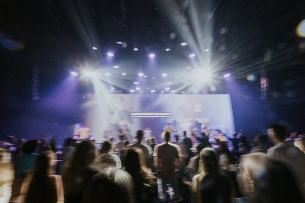 People stand in front of a stage with musicians and instruments, colorful lights shine.