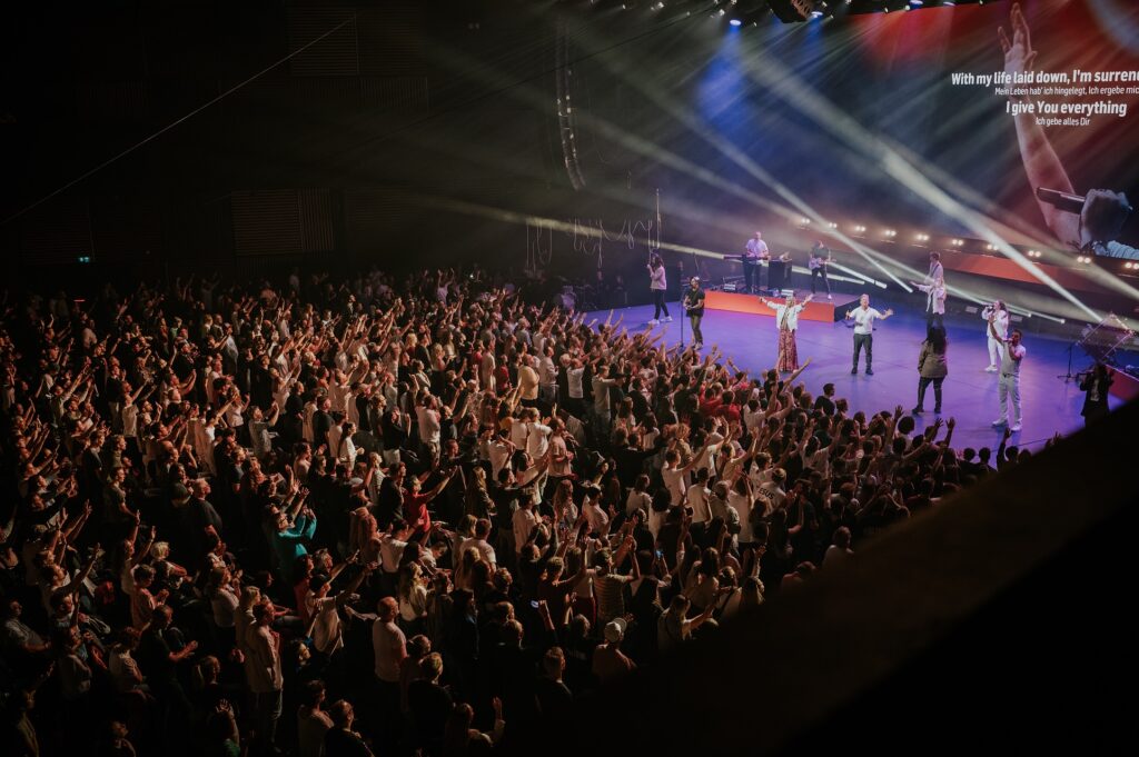 Large audience in front of a stage with several singers and musicians, stage lighting.