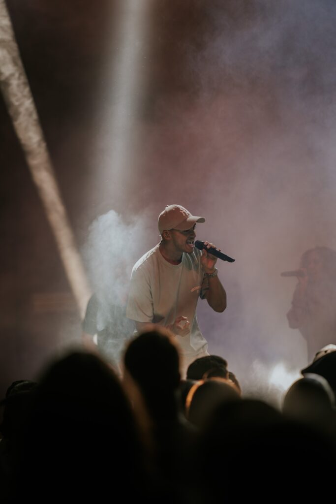 A man wearing a beige cap and white T-shirt holds a microphone and sings.