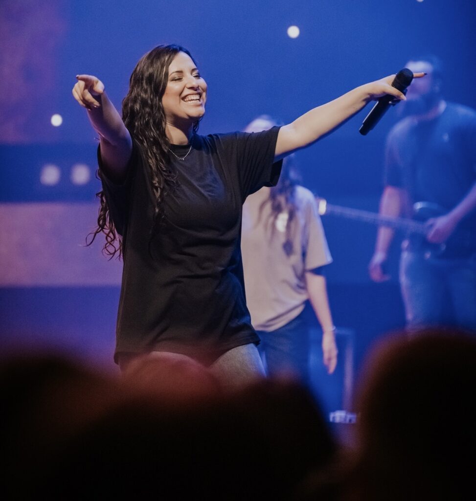 Woman with dark, wavy hair sings with microphone in hand, smiles and raises her arms.