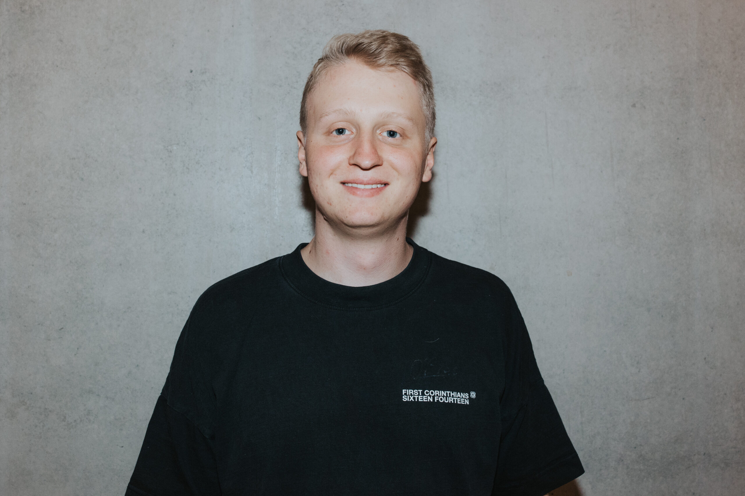 A young man with blond hair and a colorful T-shirt standing in front of a gray wall.