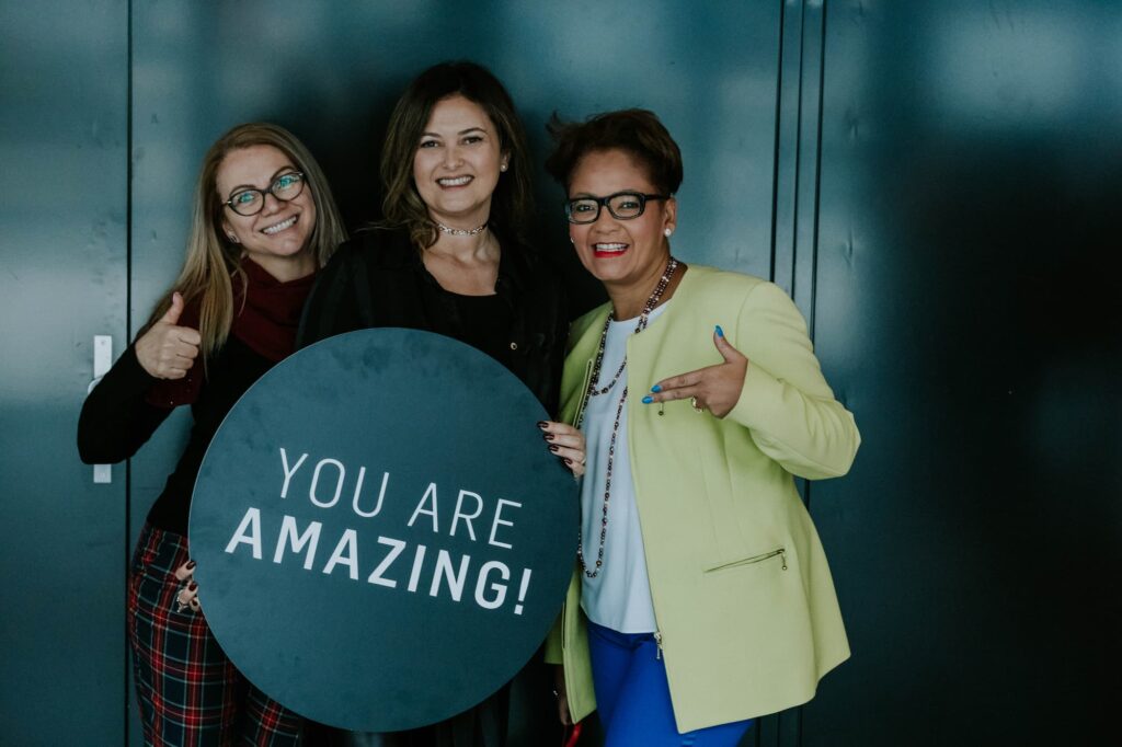 Three women hold up a sign that says “You are Amazing!”, one woman points to the sign, everyone smiles.
