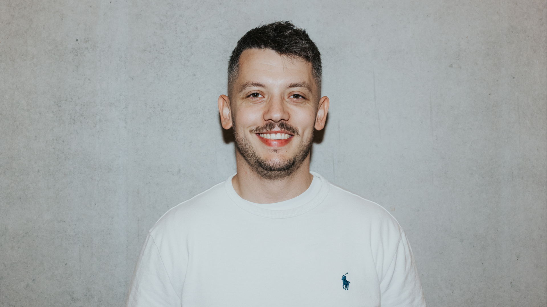 A man in a white T-shirt with a polo logo is standing in front of a gray wall