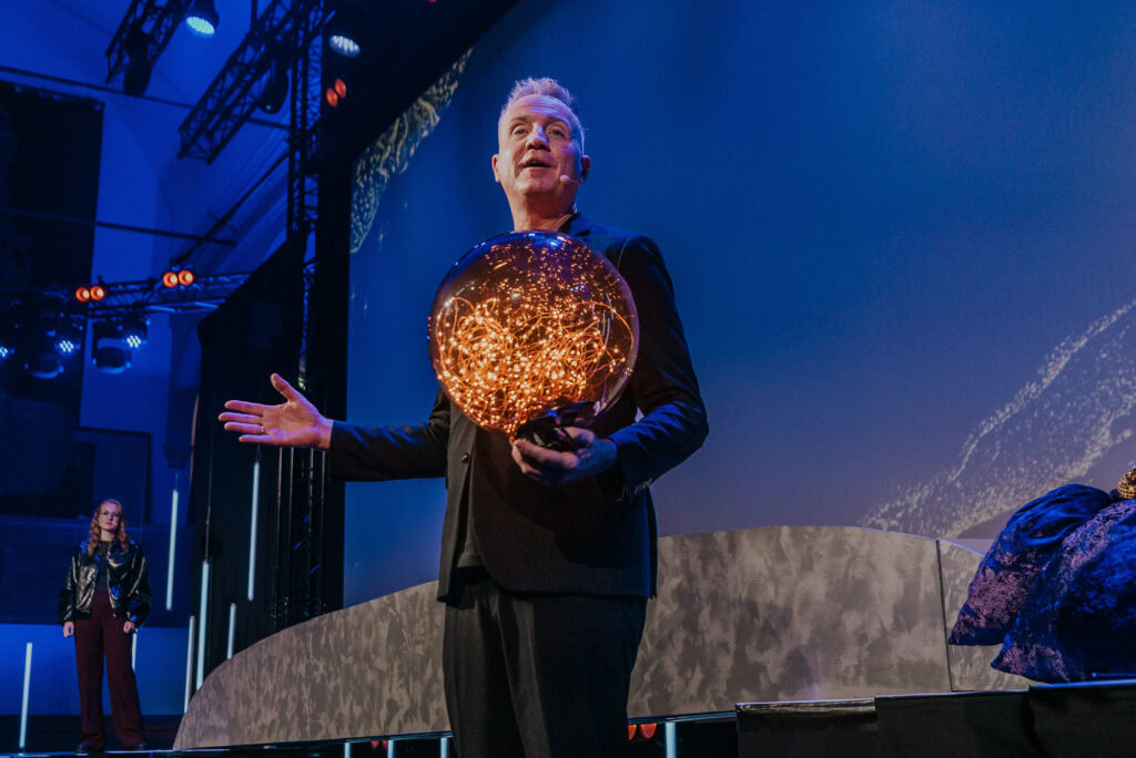 Een man houdt een bol van lichtjes vast terwijl hij naar de zaal kijkt, op een podium met een blauw-.